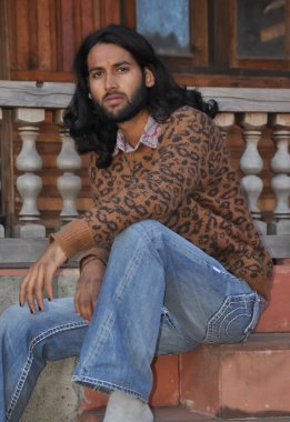 Çekici, uzun saçlı, genç bir Hintli. Tapınak merdivenlerinde elleri dizinde otururken kameraya bakıyor.