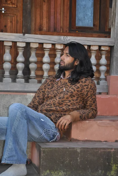 Side view of a good looking Indian young guy with long hair style, looking sideways while sitting on temple stairs