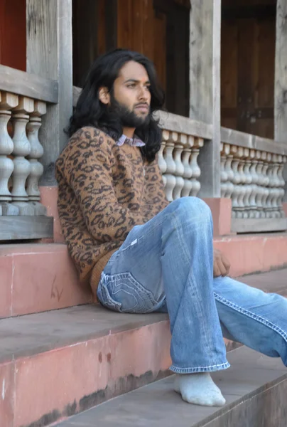 Selective focus side view of a attractive Indian young guy with long hair, looking sideways while sitting in temple stairs