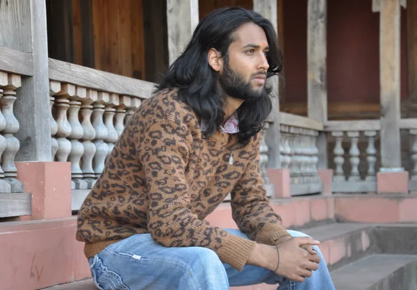 Side view of a good looking long haired Indian young guy, looking sideways while sitting in temple stairs with leaning forward