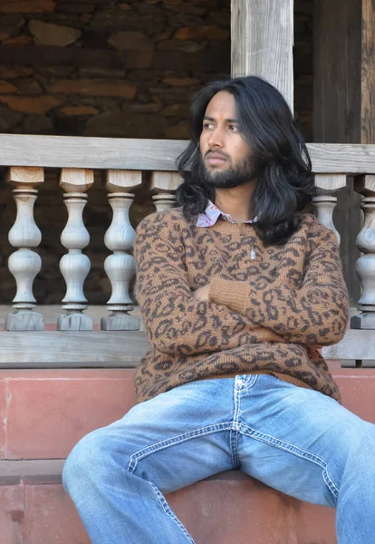 A long haired Indian young guy posing with crossed arms, looking sideways while sitting on temple stairs