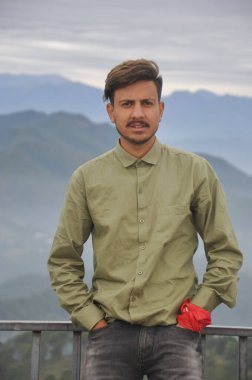 A charming Indian young guy looking at camera with hands in pocket while standing against the background of mountains