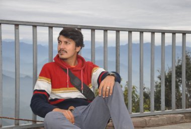 A young guy looking sideways, sitting by safety barrier with the background of mountains