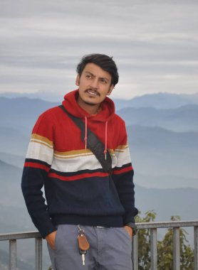 A good looking young guy looking up, with hands in pocket while leaning against safety barrier against the background of mountains
