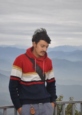 A young guy looking down, with hands in pocket while leaning against safety barrier against the background of mountains