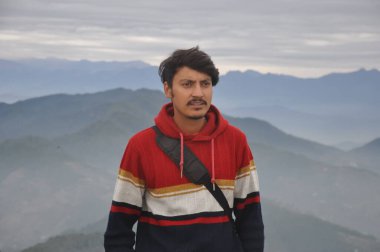 Waist-up of a handsome Indian young guy looking sideways while standing against the background of mountains 
