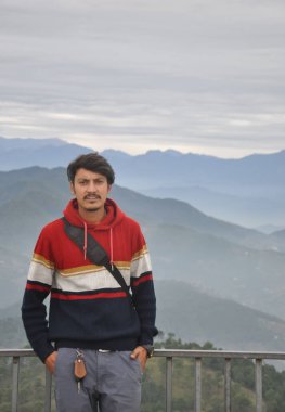 Front view of a young guy looking at camera, posing with hands in pocket while standing against the background of mountains