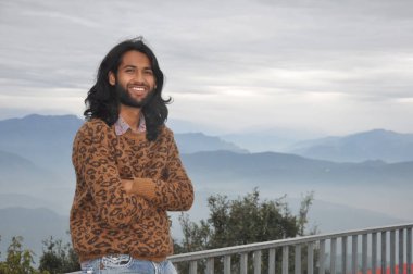 A smiling Indian young man with long hair and beard looking at camera while posing with crossed arms, sitting on safety barrier against the background of mountains
