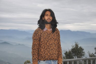A good looking Indian young man with long hair and beard looking at camera while standing against the background of mountains