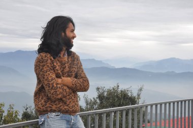A smiling long haired and bearded Indian young man looking sideways while posing with crossed arms, sitting on safety barrier against the background of mountains