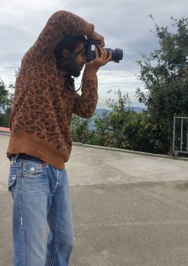 A charming Indian young man taking photo with DSLR camera while standing outdoors 
