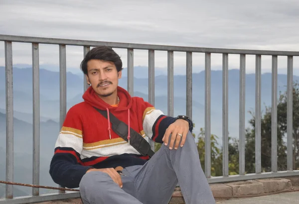 A handsome Indian young guy looking at camera, sitting by safety barrier with the background of mountains