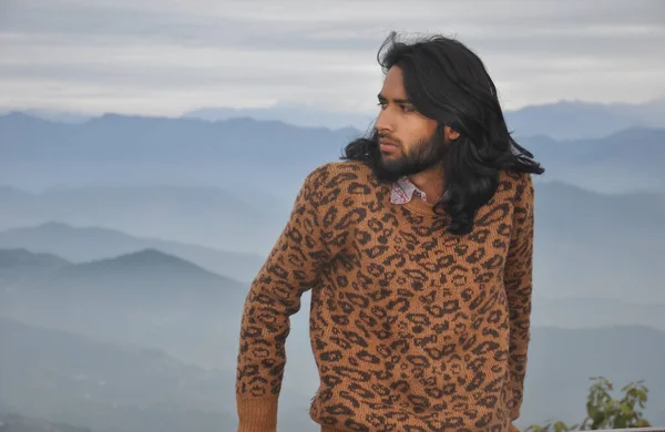 A long haired and bearded Indian young man looking sideways while standing against the background of mountains