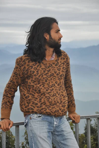 A sexy long haired and bearded Indian young man with looking sideways while sitting on safety barrier against the background of mountains