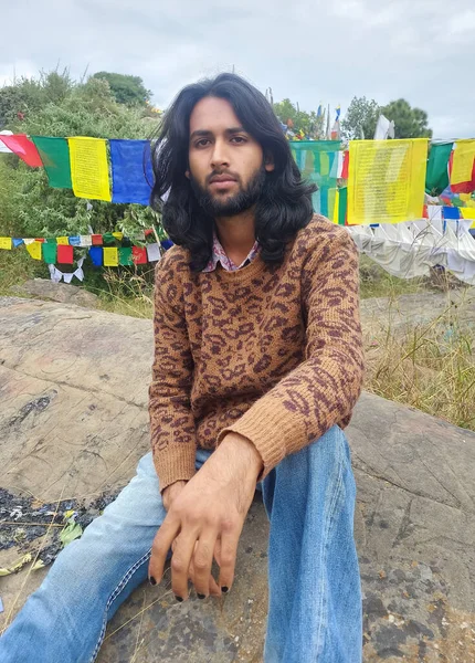 A young man with long hair and beard looking at camera while sitting over rock with the background of Tibetan prayer flags in Rewalsar