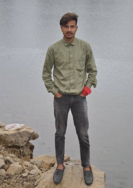 Full length of a young guy looking at camera, posing with hands in pocket while standing on stone by the lake