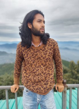 Front view of a Indian young guy with long hair and beard looking sideways while standing with leaning on safety barrier against the background of mountains