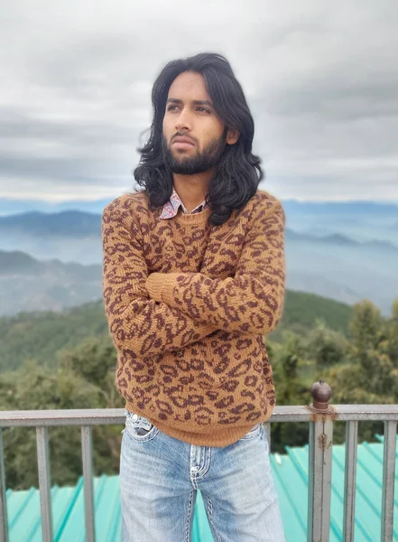 Front view of a south asian young guy with long hair and beard looking sideways while posing with crossed arms, standing with leaning on safety barrier against the background of mountains