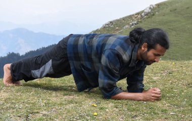 Full length of a handsome Indian young man with ponytail hair doing plank exercise in the top of mountain