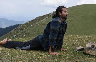 Full length of a long haired handsome young Indian man doing Bhujangasana (Ashtanga) Cobra Yoga Pose in the mountain 