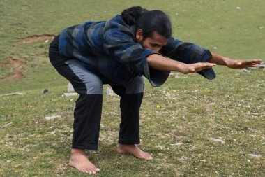 A long haired Indian young man practicing Bear Pose (Chair Pose Variation Forward Stretch) in outdoor. A young guy doing Utkatasana yoga pose (Variation Forward Stretch).