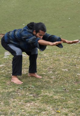 A long haired Indian young man practicing Bear Pose (Chair Pose Variation Forward Stretch) in outdoor. A young guy doing Utkatasana yoga pose (Variation Forward Stretch).