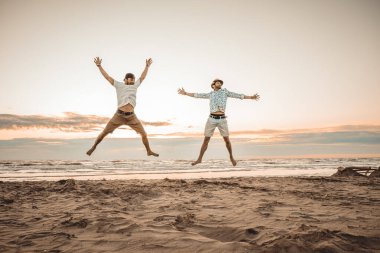 Mutlu arkadaşlar yaz tatillerini gün batımında kumsalda geçiriyorlar - iki çocuk güneşin doğuşunda plajda zıplıyor - tatil kavramı