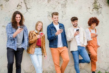 Dışarıda cep telefonu kullanan çok kültürlü bir arkadaş grubu internete girip duvarın yanında duran akıllı telefonları tutan öğrenciler.