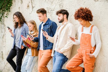 Dışarıda cep telefonu kullanan çok kültürlü bir arkadaş grubu internete girip duvarın yanında duran akıllı telefonları tutan öğrenciler.