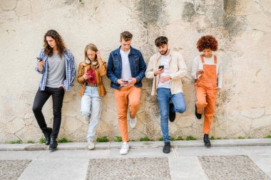 Dışarıda cep telefonu kullanan çok kültürlü bir arkadaş grubu internete girip duvarın yanında duran akıllı telefonları tutan öğrenciler.