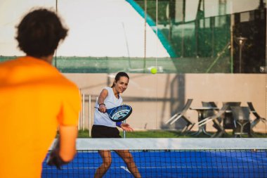 Genç öğretmen, öğrencisine Padel dersi verirken gözetliyor. Koç, kıza tenis kortunda nasıl Padel oynanacağını öğretiyor.
