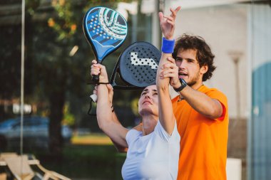 Genç öğretmen, öğrencisine Padel dersi verirken gözetliyor. Koç, kıza tenis kortunda nasıl Padel oynanacağını öğretiyor.