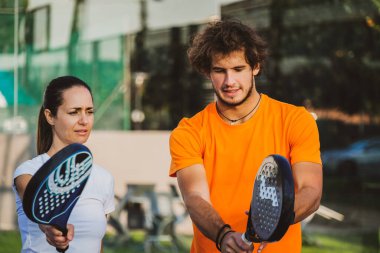 Genç öğretmen, öğrencisine Padel dersi verirken gözetliyor. Koç, kıza tenis kortunda nasıl Padel oynanacağını öğretiyor.