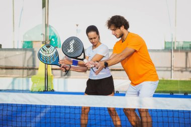 Genç öğretmen, öğrencisine Padel dersi verirken gözetliyor. Koç, kıza tenis kortunda nasıl Padel oynanacağını öğretiyor.