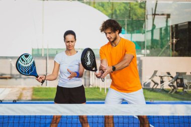 Genç öğretmen, öğrencisine Padel dersi verirken gözetliyor. Koç, kıza tenis kortunda nasıl Padel oynanacağını öğretiyor.