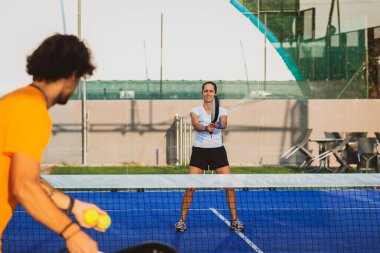 Genç öğretmen, öğrencisine Padel dersi verirken gözetliyor. Koç, kıza tenis kortunda nasıl Padel oynanacağını öğretiyor.
