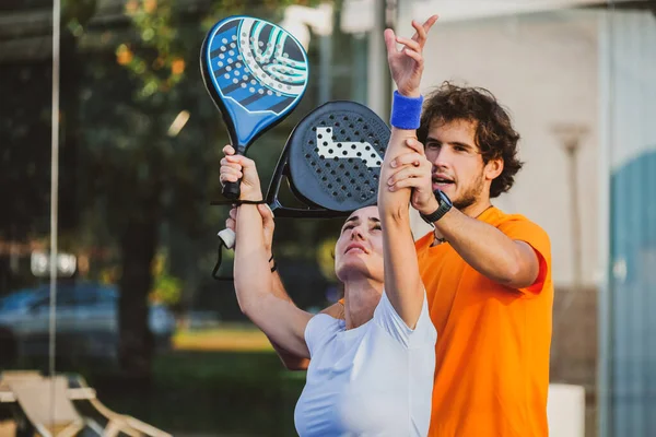 Genç öğretmen, öğrencisine Padel dersi verirken gözetliyor. Koç, kıza tenis kortunda nasıl Padel oynanacağını öğretiyor.
