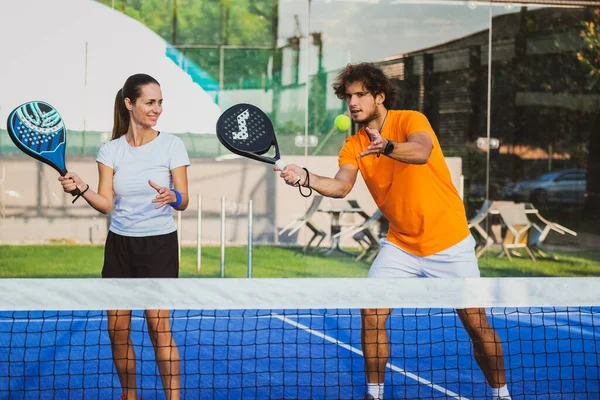 Genç öğretmen, öğrencisine Padel dersi verirken gözetliyor. Koç, kıza tenis kortunda nasıl Padel oynanacağını öğretiyor.