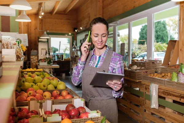 Bir markette dijital tablet kullanan ve envanter alırken cep telefonuyla konuşan mutlu kadın satış asistanının portresi dijital tabletle çevrimiçi market siparişi hazırlıyor.