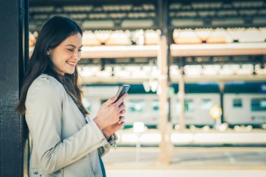 Bir tren istasyonunda beklerken akıllı telefon kullanan gülümseyen iş kadını, tren istasyonunda beklerken cep telefonu kullanan çekici bir genç kız.