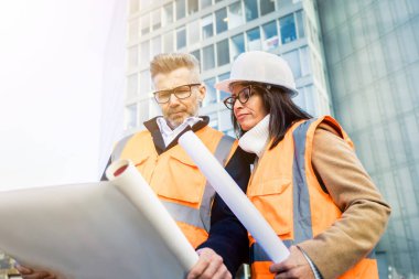 Turuncu iş yeleği ve miğfer giyen iki mühendis meslektaşı inşaat belgelerini inceliyor. Takım konsepti