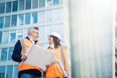 Turuncu iş yeleği ve miğfer giyen iki mühendis meslektaşı inşaat belgelerini inceliyor. Takım konsepti