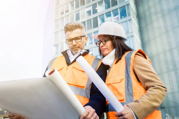 Turuncu iş yeleği ve miğfer giyen iki mühendis meslektaşı inşaat belgelerini inceliyor. Takım konsepti
