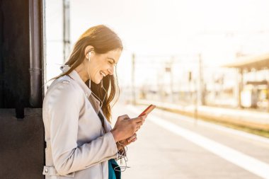 Bir tren istasyonunda beklerken akıllı telefon kullanan gülümseyen iş kadını, tren istasyonunda beklerken cep telefonu kullanan çekici bir genç kız.