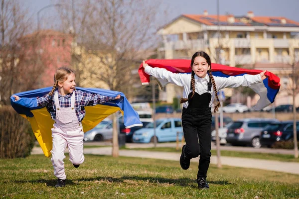 公園で幸せを実行しているウクライナとロシアのフラグを持つ美しい小さな女の子 ウクライナ人とロシア人の平和の概念 ストック写真