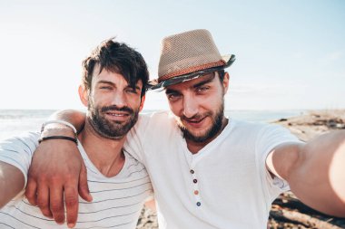 İki genç erkek arkadaş gün batımında sahilde birbirlerine sarılıp selfie çekiyorlar. Arkadaşlık ve aşk kavramı