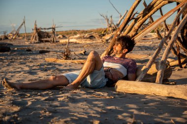 Yaz şapkasıyla gevşeyen genç bir adam çöl sahilindeki tahta kütüğün üzerinde gün batımına bakan bir birayla yatıyordu.
