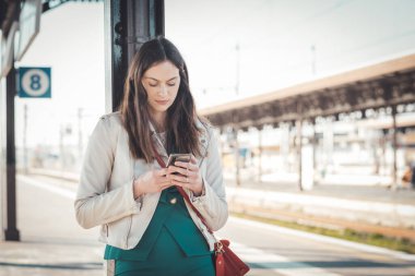 Bir tren istasyonunda beklerken akıllı telefon kullanan gülümseyen iş kadını, tren istasyonunda beklerken cep telefonu kullanan çekici bir genç kız.