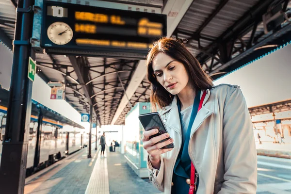 Young woman with departure times behind her waiting for her train while holding her mobile phone - Woman looking at the clock in the train station while her train is delayed - Transportation and urban life concept