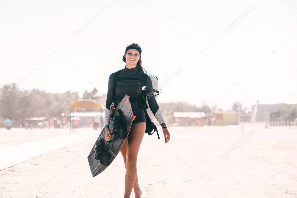 Portrait of beautiful smiling girl posing with a surfboard on the beach and wearing kitesurfing equipment - concept of sporty woman and summer sport activity